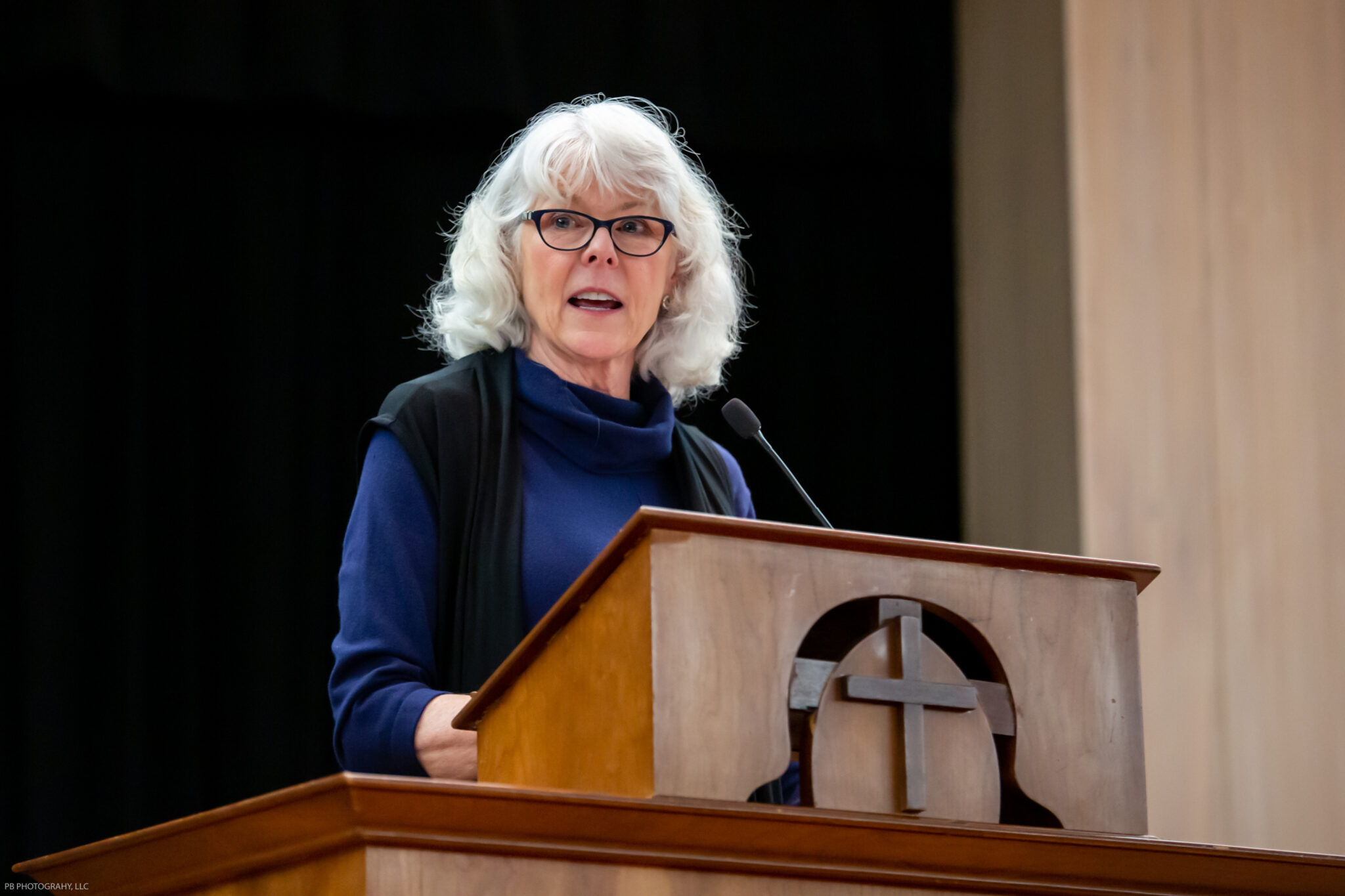 A white woman with white hair and a blue sweater behind a wooden lectern