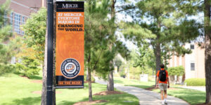 A student in an orange shirt walking down the sidewalk with a backpack on. In the foreground, a banner reads, "At Mercer, everyone majors in changing the world."