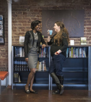 One black and one white woman talking while leaning against a shelf and drinking from mugs