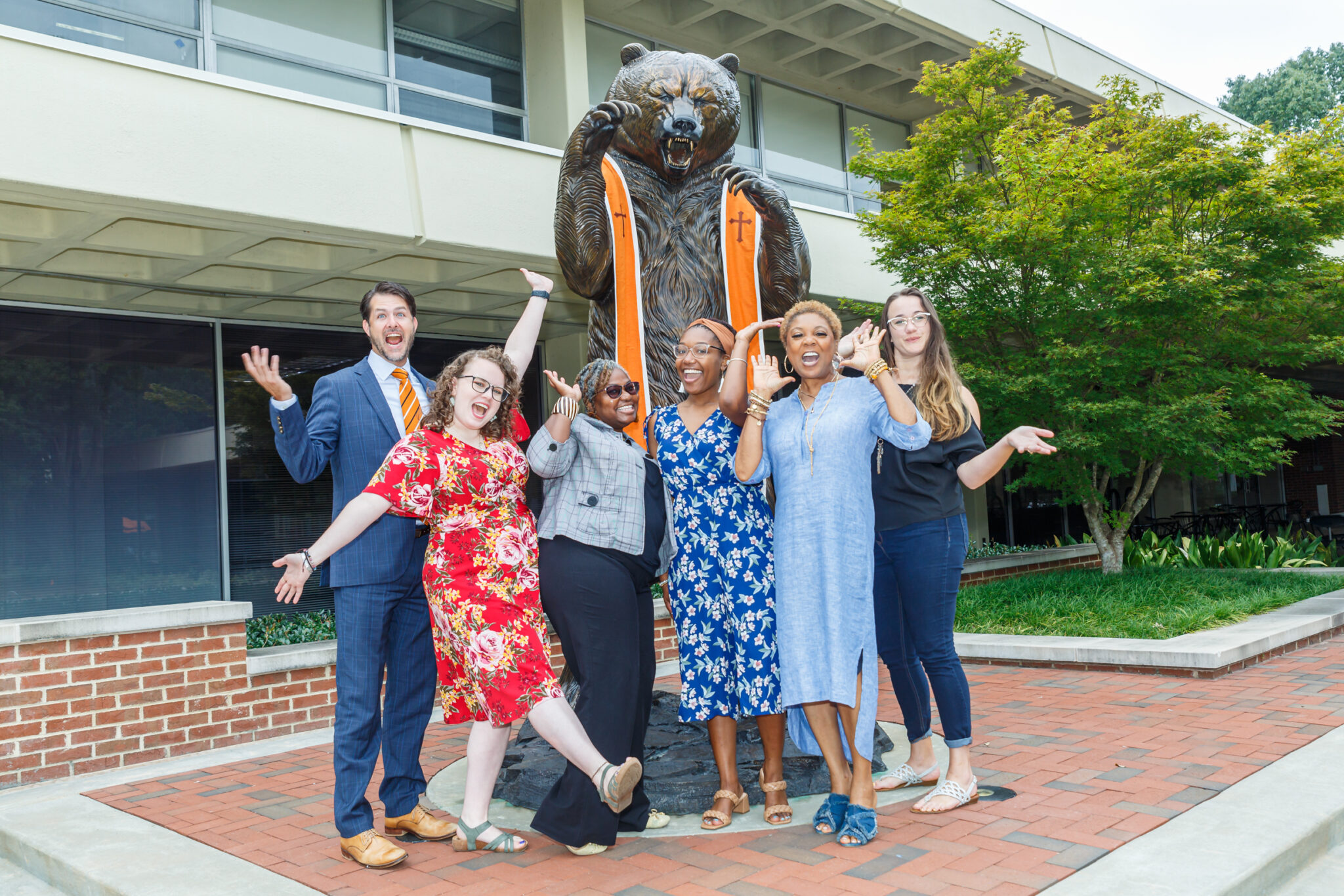 Six individuals of mixed race and gender celebrating in front of a large bear statue