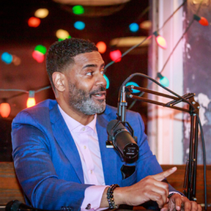 Black man in a blue suite speaking into a microphone recording a podcast