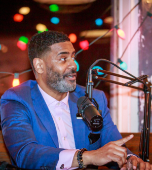 Black man in a blue suite speaking into a microphone recording a podcast