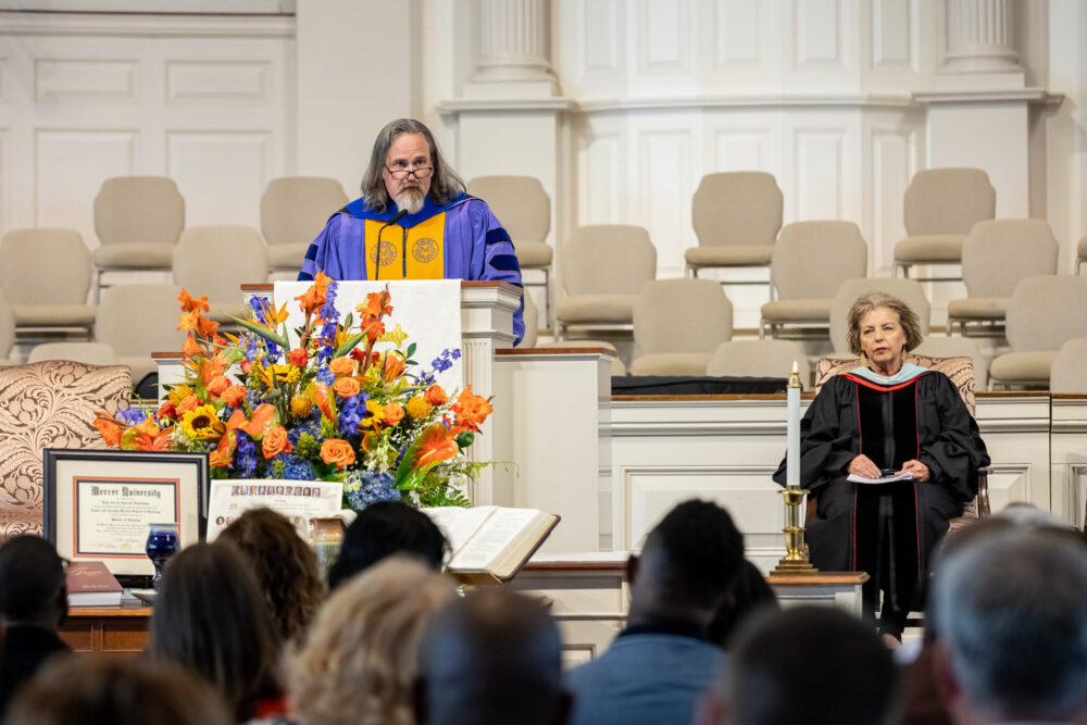White male in blue and gold regalia speaking from behind a pulpit