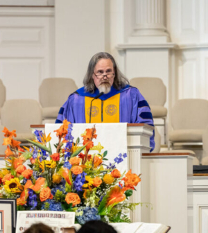 White male in blue and gold regalia speaking from behind a pulpit