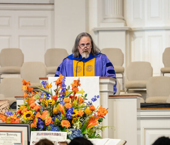 White male in blue and gold regalia speaking from behind a pulpit