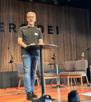 White male in green t-shirt and jeans speaking on a stage