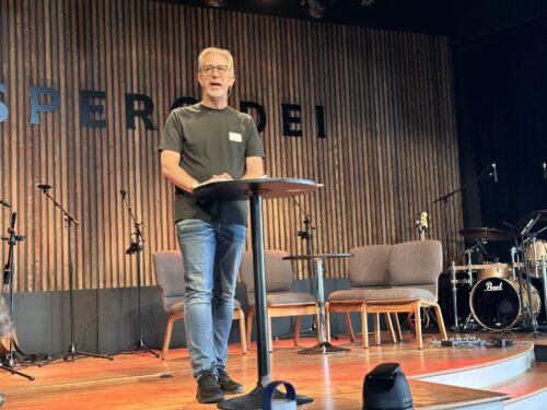 White male in green t-shirt and jeans speaking on a stage